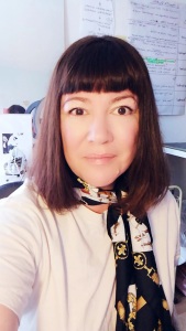 White woman, Andree Girard-Kemp, fashion design expert with a brown bob hairstyle and blunt fringe staring at the camera and smiling. Wearing a cream t-shirt and vintage silk neck scarf